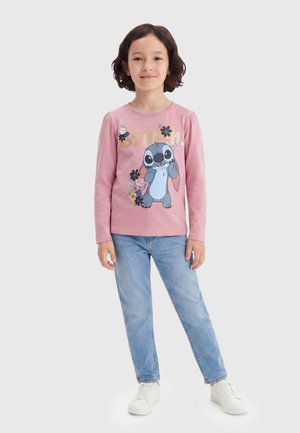 Enfant aux cheveux bouclés portant un t-shirt rose à manches longues avec un personnage Stitch et des fleurs, un jean bleu clair et des baskets blanches.