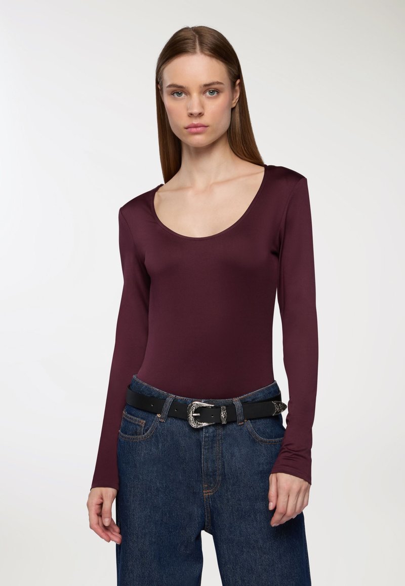 Body a maniche lunghe bordeaux con scollo rotondo, abbinato a jeans in denim blu a vita alta e una cintura nera con fibbia metallica.