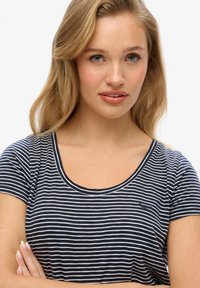 Lyshåret kvinde med lyseblå øjne iført en marineblå og hvidstribet t-shirt, krydsede arme, kigger direkte ind i kameraet.