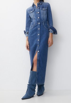 Femme portant une longue robe en jean boutonnée avec une fente à l'avant et des manches retroussées, associée à des bottes en jean pointues et décontractées.