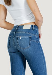 Siniset farkut, joissa on kapea malli ja takataskut, vaaleanbeige brändilogo vyötäröllä sekä ompelukoristeet.