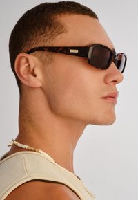 Άνδρας που φοράει γυαλιά ηλίου με σχέδιο χελώνας και κολιέ με χάντρες, εμφανιζόμενος σε κοντινό προφίλ με ουδέτερη έκφραση σε ανοιχτό φόντο.