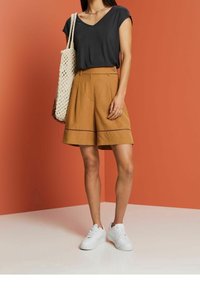 T-shirt grigio scuro a V, pantaloni corti color cammello con pieghe frontali e dettagli a trim, sneakers bianche e una borsa a tracolla intrecciata color beige.