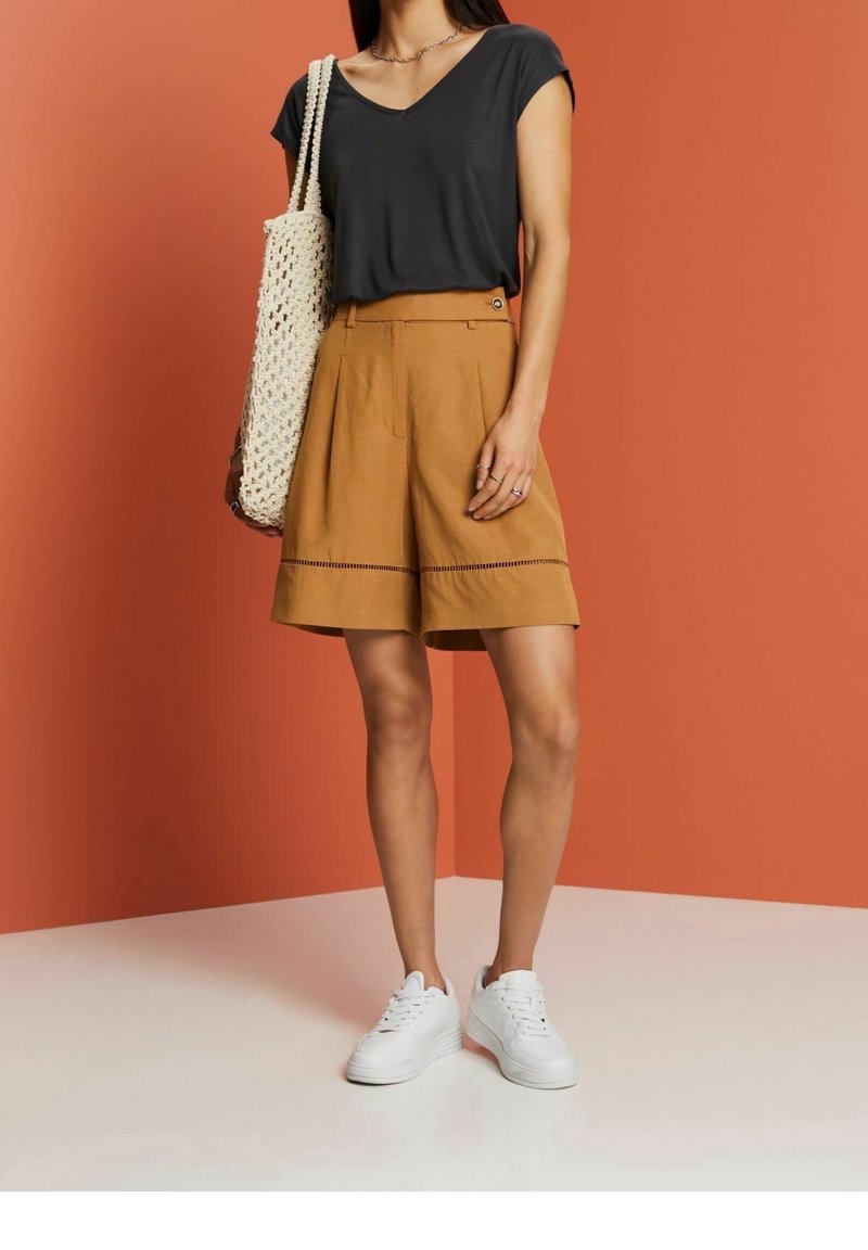 T-shirt grigio scuro a V, pantaloni corti color cammello con pieghe frontali e dettagli a trim, sneakers bianche e una borsa a tracolla intrecciata color beige.