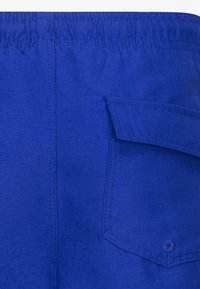 Blå stoffshorts har en elastisk midje, sidelomme med klaff og glatt tekstur. Søm-detaljer fremhever designet.