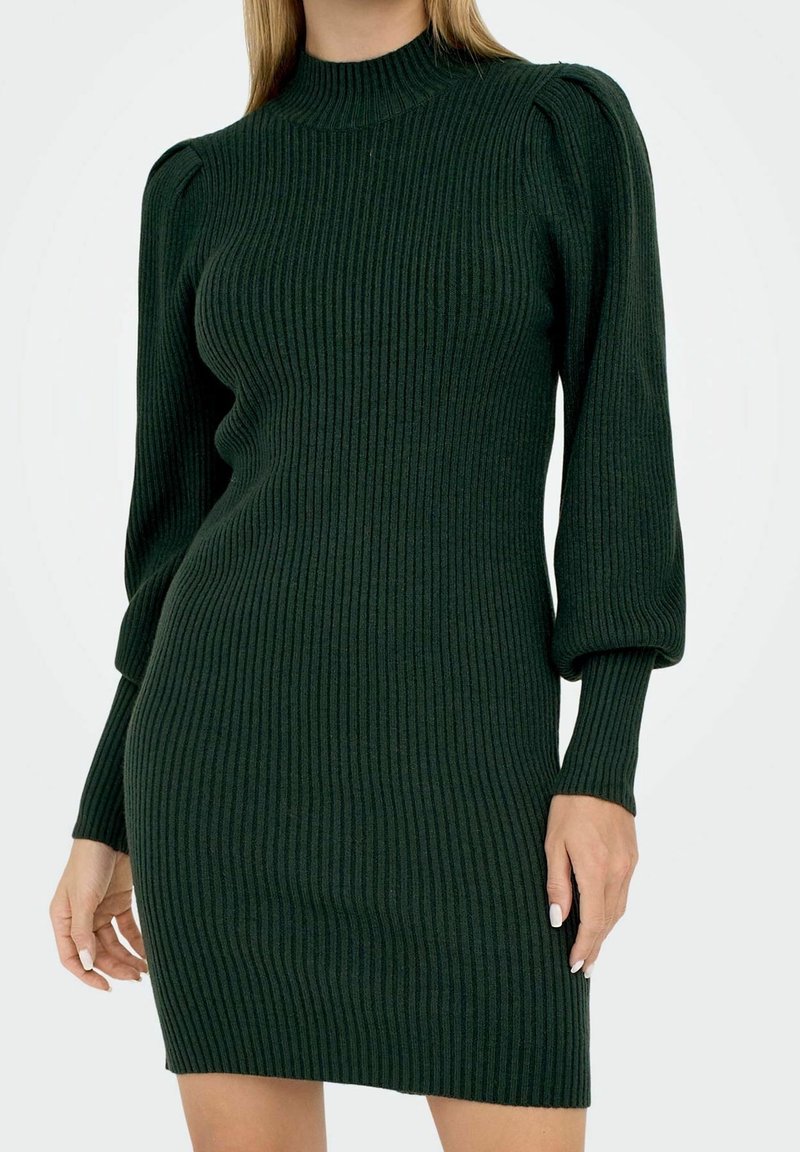 Femme portant une robe en maille côtelée vert foncé, moulante, à manches longues et col montant, debout devant un fond clair uni.