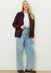 Bordeaux ritsjack over een witte t-shirt, gecombineerd met lichtblauwe high-waisted jeans. Gladde stof, relaxte pasvorm en twee voorkruiszakken.