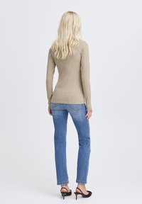 Frau mit langen blonden Haaren, die ein beiges Langarmshirt, blaue Jeans und schwarze Absatzsandalen trägt, steht mit dem Rücken zu einer weißen Wand.