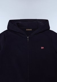 Tmavomodrá mikina na zips s mäkkou textúrou a veľkou kapucňou. Na hrudi má malý vyšívaný detail vlajky.