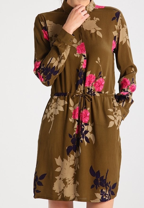 Robe marron à manches longues avec des motifs floraux roses et beiges, col montant, boutons sur le devant et ceinture à la taille, portée avec une main près du visage.