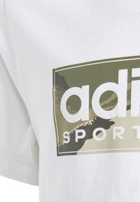 Camiseta de algodón blanca con un gran logo de camuflaje en verde y beige que incluye el texto "adidas SPORT" en letras blancas en negrita. Mangas cortas.