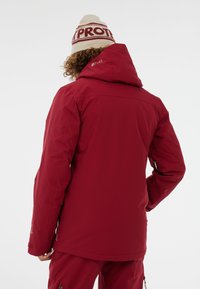 Rode waterdichte jas met een capuchon, met een gladde textuur en zichtbare branding op de achterkant. Gecombineerd met bijpassende broek.