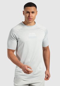 Jasnoszara koszulka sportowa z krótkim rękawem, wykonana z teksturowanej tkaniny, z okrągłym dekoltem. Posiada logo na klatce piersiowej w nieco ciemniejszym odcieniu szarości.