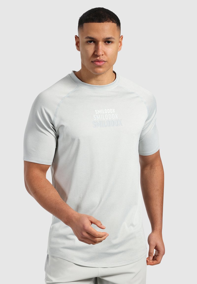 Jasnoszara koszulka sportowa z krótkim rękawem, wykonana z teksturowanej tkaniny, z okrągłym dekoltem. Posiada logo na klatce piersiowej w nieco ciemniejszym odcieniu szarości.