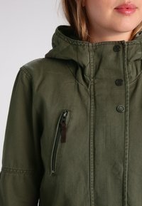 Olivově zelená bunda s kapucí, s kapsou na zip a cvoky. Vyrobena z měkké tkaniny s hladkým povrchem.