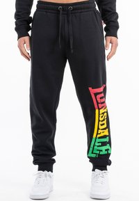 Pantalones de chándal de algodón negro con un ajuste tapered, que presentan un colorido logo de “Lonsdale” en rojo, amarillo y verde a lo largo de la pierna izquierda. Cintura ajustable.