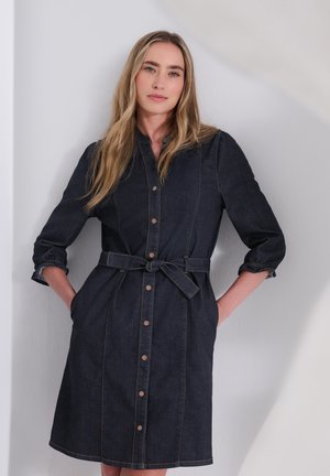 Femme aux longs cheveux blonds portant une robe en jean foncé ceinturée avec des boutons, debout les mains dans les poches contre un mur clair uni.