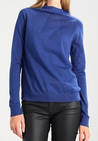 Donna che indossa un maglione blu a maniche lunghe con scollo rotondo abbinato a pantaloni neri aderenti su uno sfondo neutro.