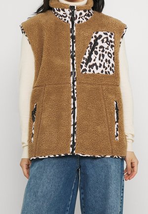 Brązowa bezrękawnikowa kamizelka sherpa z akcentami zamka błyskawicznego w panterkę, noszona na kremowym swetrze i niebieskich dżinsach, widok z przodu.