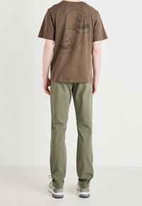 T-shirt di cotone marrone con grafica di teschio nera sulla schiena, abbinato a pantaloni verdi e sneaker grigie, visto da dietro.