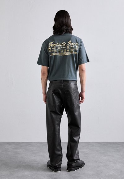 Camiseta de algodón gris con texto gráfico dorado en la parte posterior, combinada con pantalones de cuero negros. Destacan las texturas suaves y el ajuste relajado.