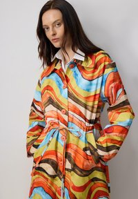 Palton colorat cu modele featuring designuri ondulate în nuanțe de portocaliu, albastru, galben și maroniu. Are talie cu curea, închidere cu nasturi pe față și mâneci largi.
