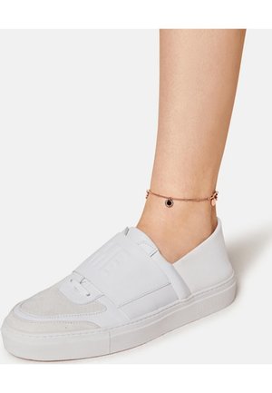 Jambe inférieure nue portant un bracelet de cheville fin en or rose avec un pendentif noir et une sneaker slip-on blanche à semelle épaisse.