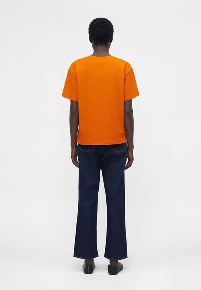 Άτομο που στέκεται με την πλάτη γυρισμένη, φορά φωτεινό πορτοκαλί κοντομάνικο πουκάμισο, σκούρα μπλε τζιν και μαύρα παπούτσια σε απλό φόντο.