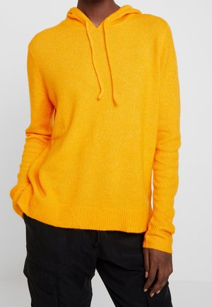 Modèle portant un pull en maille jaune vif à capuche et à cordons, ainsi qu’un pantalon noir, sur un fond gris clair.