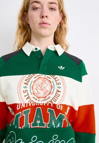 Zaļš un balts polo krekls ar Miami universitātes grafiku, sarkaniem un oranžiem svītrainiem, kā arī mazu Adidas logo uz krūšu.