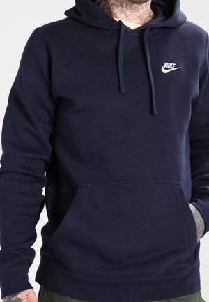 Homme portant un sweat à capuche Nike bleu marine avec poche avant et cordons, montrant des tatouages au cou et à la main.