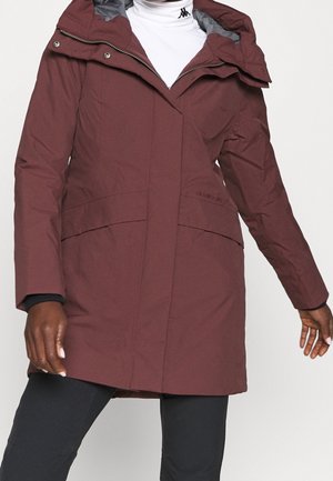 Personne portant un manteau d'hiver bordeaux arrivant aux genoux avec un grand col, une fermeture éclair à l'avant et un pantalon noir sur un fond uni.