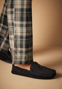 Svarta slippers i mocka med vävd textur och pälsfoder, kombinerade med rutiga byxor i grå, svart och orange toner.