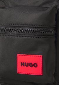 Dettaglio di uno zaino in nylon nero con un'etichetta rettangolare rossa ben visibile con la scritta "HUGO" in nero. Chiusura a zip con un tirante testurizzato.