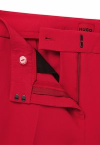 Pantaloni rossi con una texture liscia, dotati di chiusura con bottone, tasca con zip nera e etichetta con marchio. Design pulito e forma su misura.