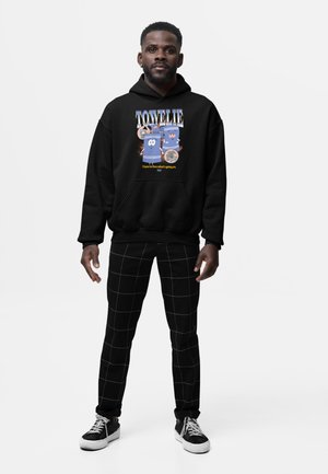 Hombre de pie, vistiendo una sudadera negra con capucha con dibujos de toallas y texto "TOWELIE" y pantalones negros con patrón de cuadrícula blanca sobre fondo gris liso.