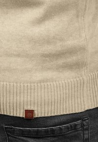 Beżowy sweter dziergany z prążkowanym dołem, z małą brązową skórzaną metką; zestawiony z ciemnoszarymi dżinsami.