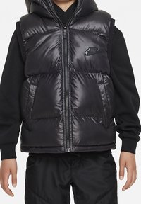 Chaleco acolchado negro con capucha, fabricado en tejido brillante. Presenta un cierre fronta y un logo de Nike negro en el pecho. Diseñado para proporcionar calidez y comodidad.