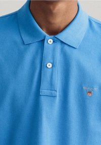 Polo bleu clair en tissu texturé, doté d'une patte à deux boutons, d'un col, et d'un petit logo GANT avec détail bouclier.