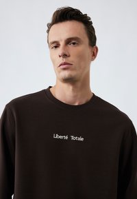 Ruskea collegepaita, jossa on valkoisella brodeerattu teksti "Liberté Totale", pyöreä kaula-aukko ja pehmeä, tekstuurinen kangas.