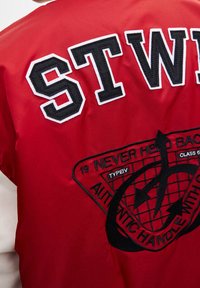 Veste bomber en satin rouge avec des accents blancs et noirs, présentant de grandes lettres "STW" et un patch brodé avec un design géométrique.