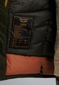 Zelená vosková parka s černou polstrovanou podšívkou a oranžovým interiérem. Obsahuje štítek se specifikacemi, zipové kapsy a nastavitelné manžety.