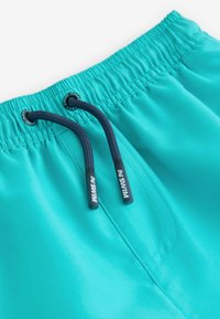 Pantaloni da bagno turchesi con cintura elastica, dotati di lacci navy e punte con marchio. Texture liscia con cuciture a sbalzo.