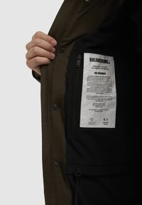 Maslinasto zelena vanjska jakna s crnim interijerom. Unutrašnjost sadrži zip džep i etiketu s otisnutim tekstom. Vidljiva prednja kopča na dugme.