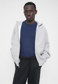 Sudadera gris con cremallera y interior blanco, llevada sobre una camiseta azul marino. Combinada con pantalones deportivos negros. Tela lisa y corte holgado.