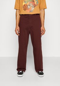 Bordeaux jeans med rak bendesign, prydda med en framficka och en knappstängning. Bärs med en brun T-shirt och svarta sneakers.