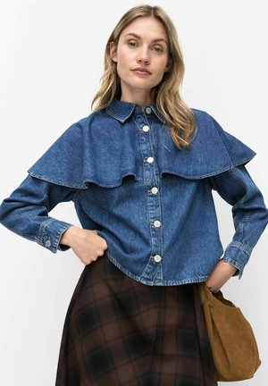 Femme portant une chemise en denim bleu à boutons avec un large col superposé et une jupe à carreaux foncés, tenant un sac en daim marron à son côté.