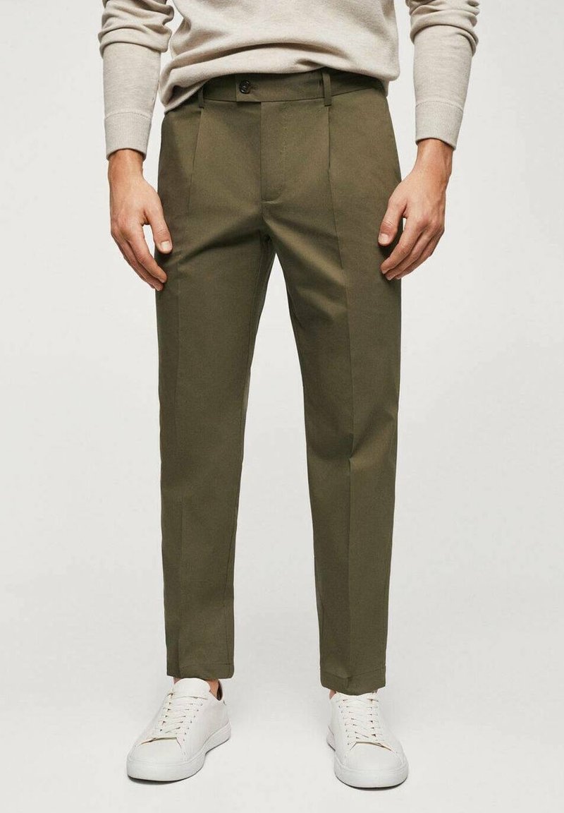 Homme portant un pantalon vert olive cintré, un pull beige et des baskets blanches, debout avec les mains détendues le long du corps.