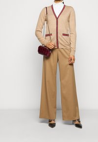 Beiger Cardigan mit V-Ausschnitt, roten und schwarzen Akzenten, kombiniert mit beigen weit geschnittenen Hosen und einer kleinen burgunderfarbenen Umhängetasche.