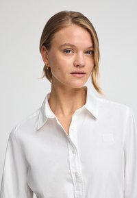 White button-up shirt with a classic collar, soft texture, and subtle branding patch. Features translucent buttons and a tailored fit.